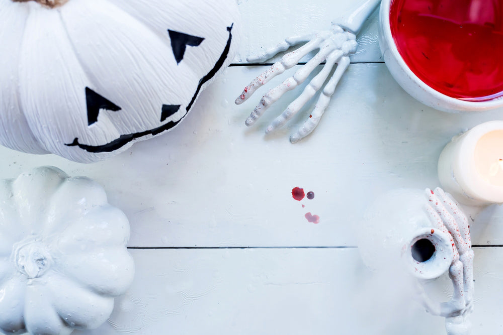 White jack-o-lantern on a white table with skeleton hands and a red drink.