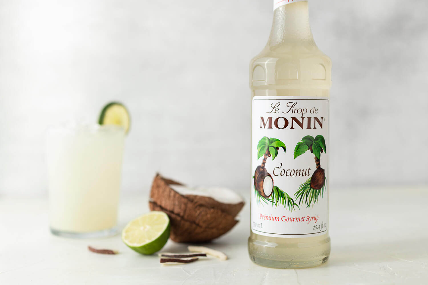 A bottle of Monin Coconut Syrup sits next to a cracked open coconut, lime slices, and a drink in a short glass