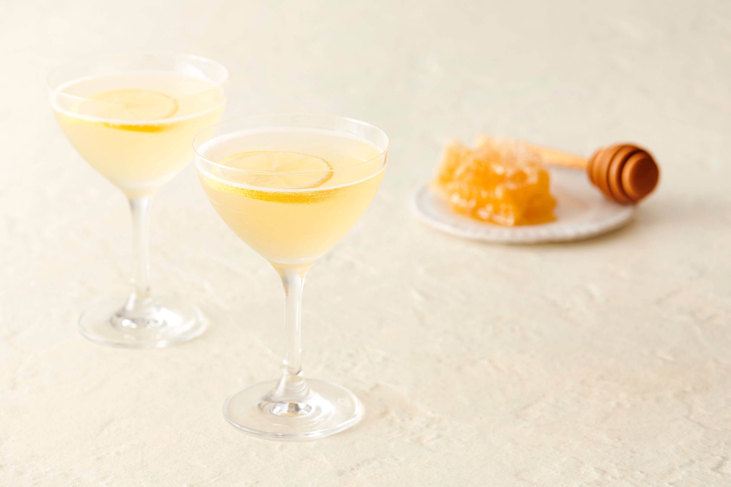 Two coupe glasses filled with light yellow liquid, with a honey comb on a plate in the background.