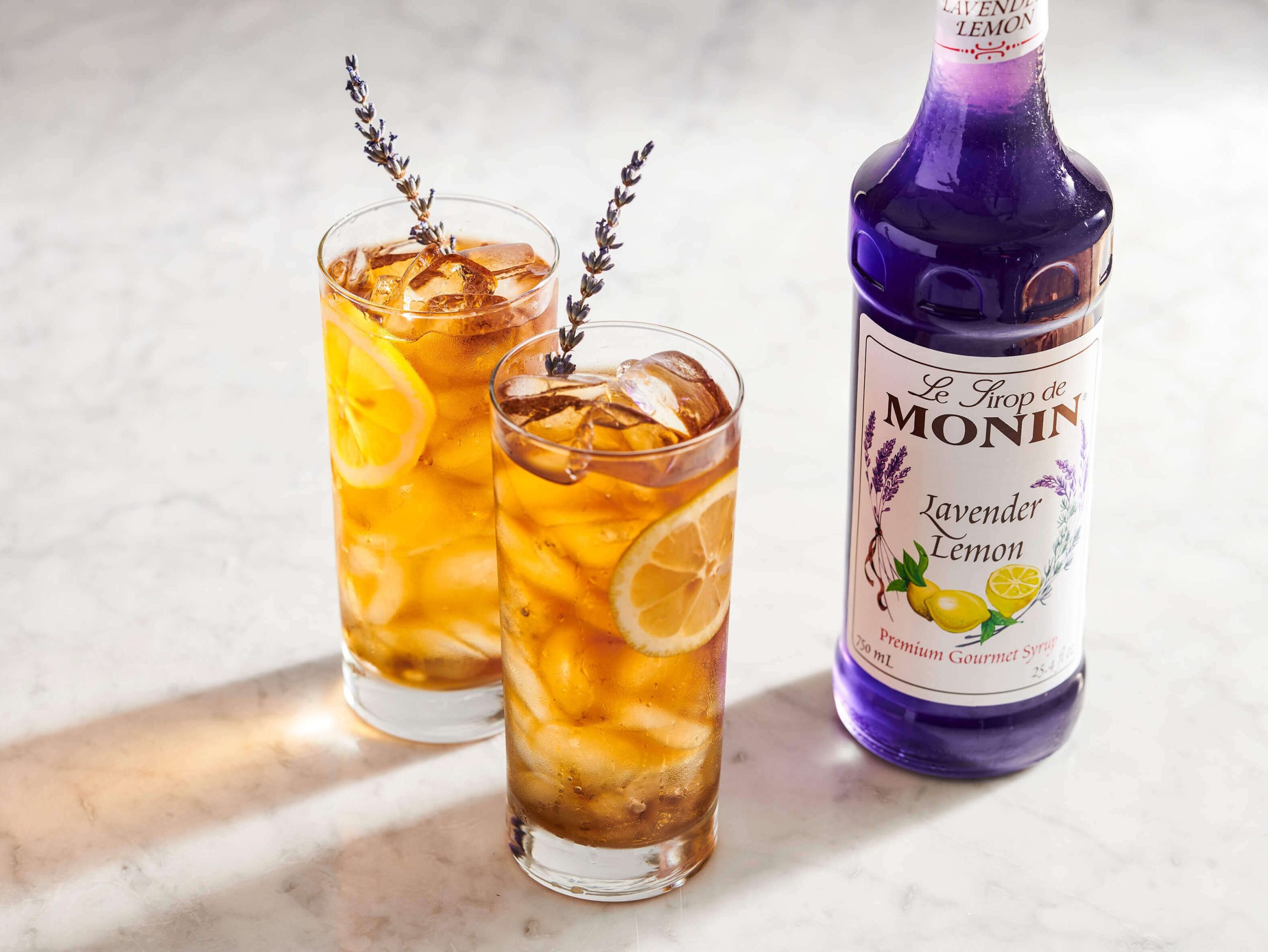 Two highball glassesj with tea beverages are placed to the left of a glass bottle of Monin Lavender Lemon syrup.