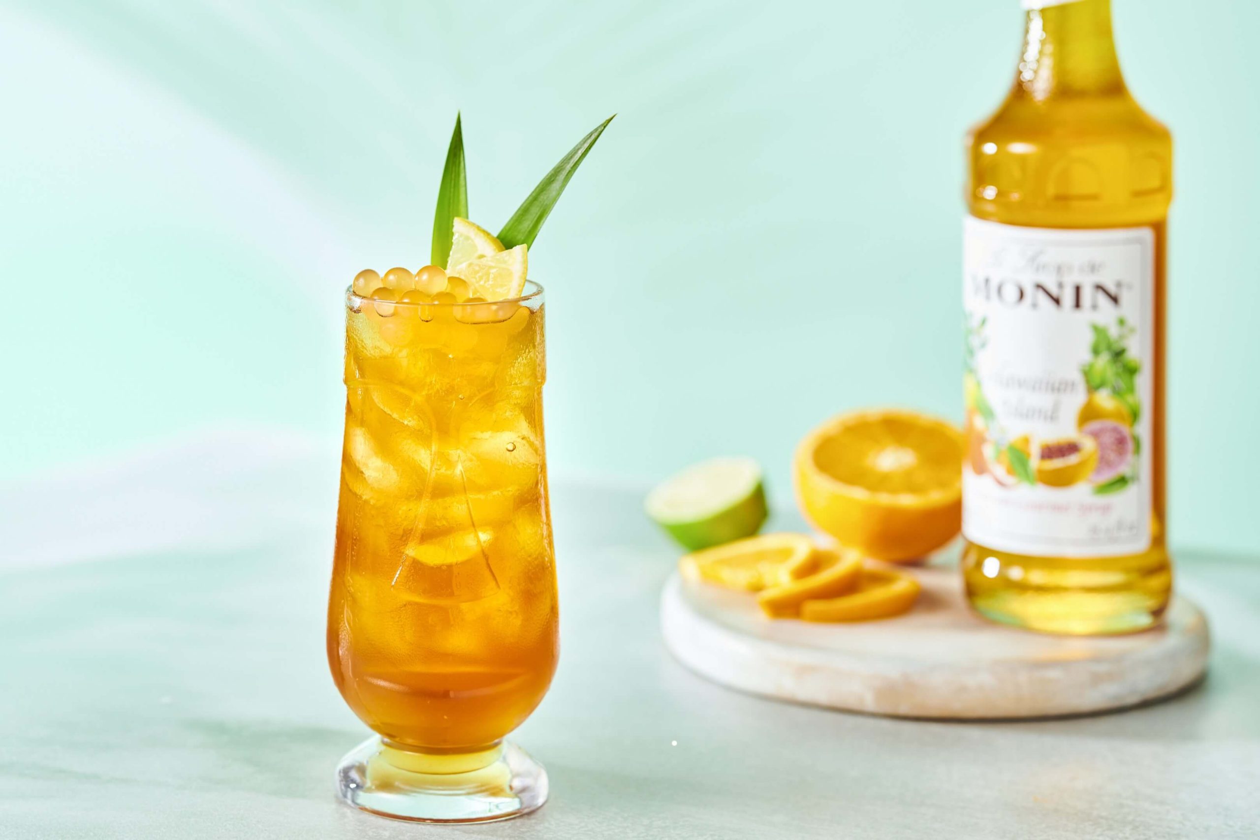 A tall glass for Hawaiian Pearl Bubble Tea, with citrus garnishes and a bottle of Hawaiian Island Syrup in the background.