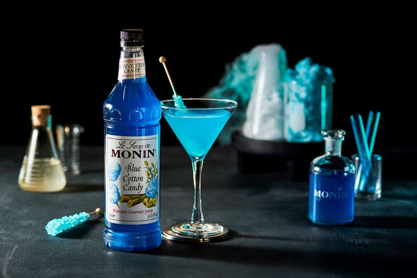 A blue Cotton Candy Martini sitting in front of a chemistry set up filled with blue liquid. All on a black backdrop.