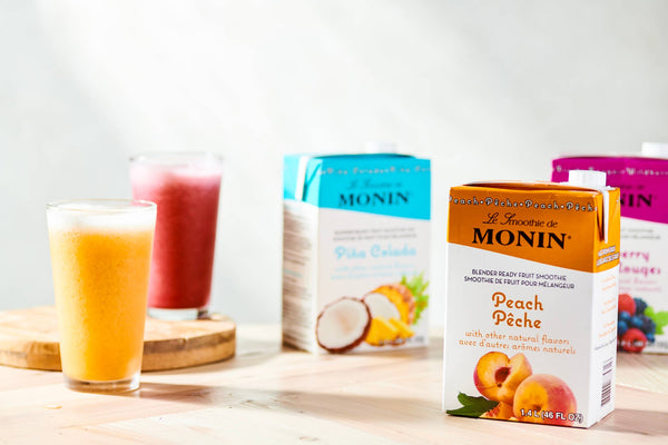 A red smoothie and yellow smoothie in large glasses next to each other. Beside them are boxes of Monin Smoothie Mixes in Peach, Pina Colada, and Wildberry