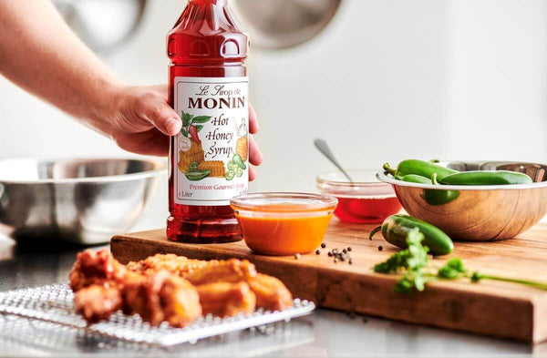 An outstretched arm setting down a bottle of Monin Hot Honey Syrup onto a cutting board. Also on the cutting board are peppers, honey in a small dish. Chicken wings blurred in the foreground.