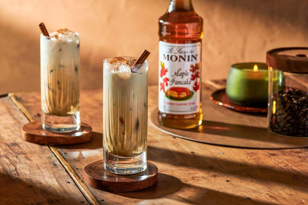 In the foreground, two iced coffees garnished with cinnamon powder and cinnamon sticks sit on a wooden table. In the background a bottle of Monni Maple Pancake Syrup, a candle, and a jar of coffee beans.