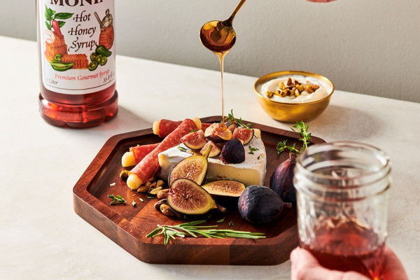 Hot Honey Syrup bottle in the background, with someone drizzling Hot Honey Syrup on a charcuterie board in the foreground.