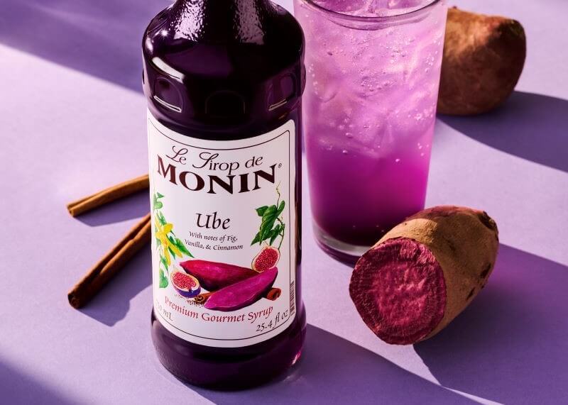 A bottle of Monin Ube Syrup against a purple background. Behind it are cinnamon sticks, an ube cut in half, and a glass of purple Ube Soda.