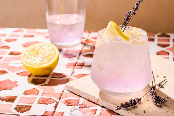 A lavender spritz in a stemless wine glass, garnished with a lavender sprig and lemon slice. IN the background is a half-empty glass cocktail shaker containing the remaining cocktail, and half a lemon sitting next to it.