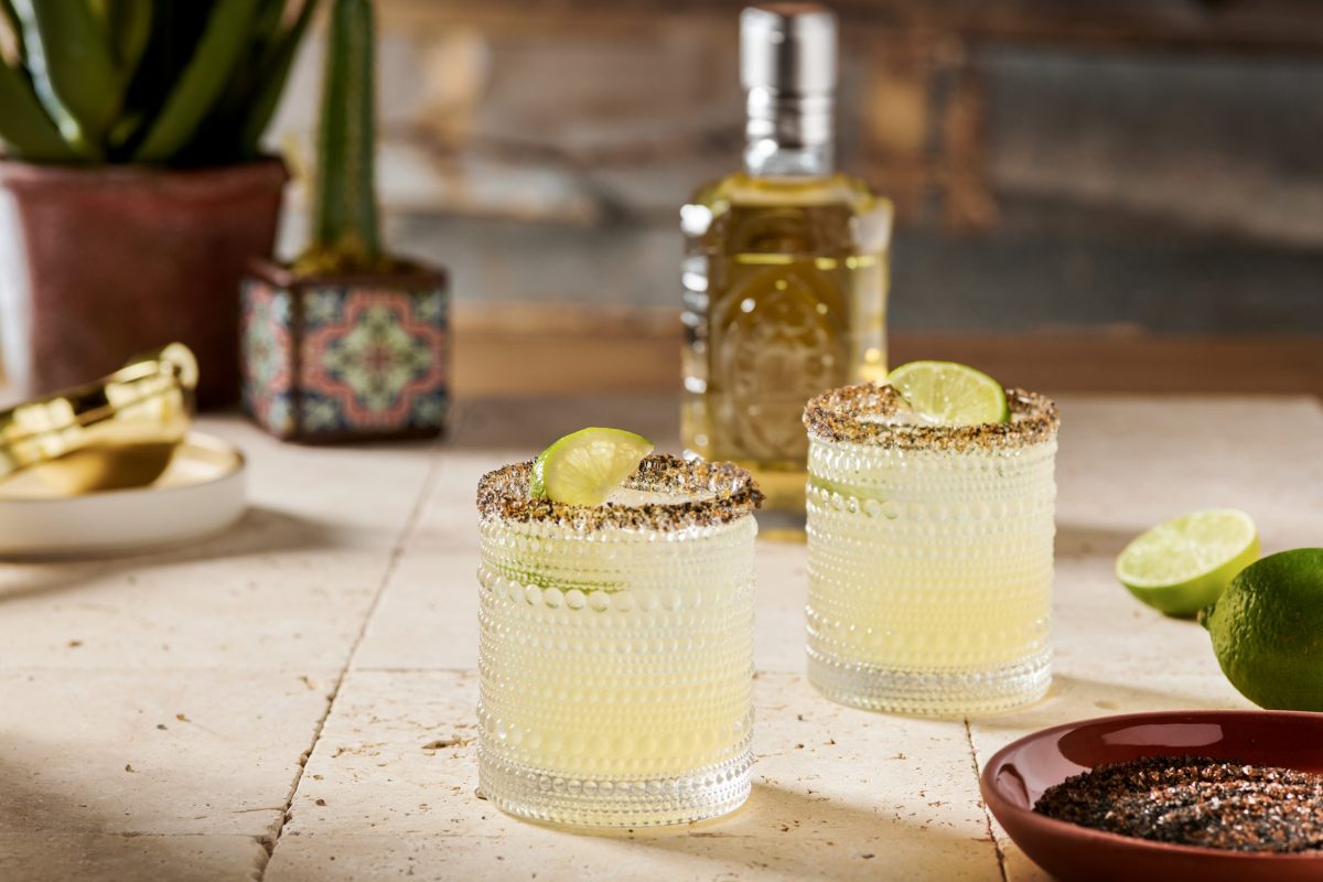 Two margarita glasses with salt rims set on a stone surface. Each has a lime garnish and there is a mezcal bottle in the background.