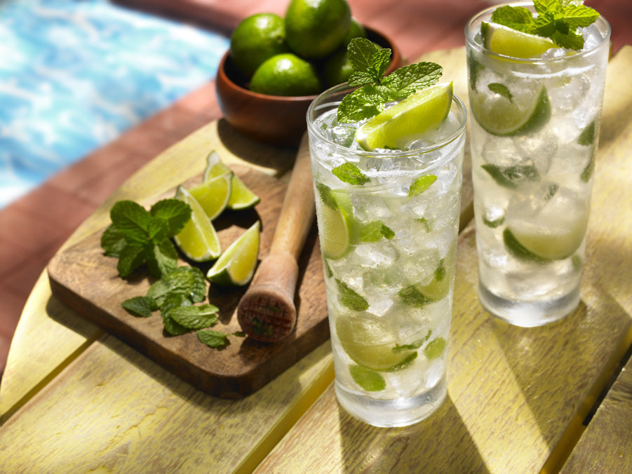 Two mojito drinks next to a muddler, lime wedges, and mint leaves.