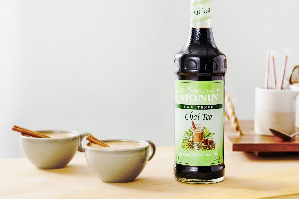 A bottle of Monin Chai Tea Concentrate sits next to two hot chai lattes on a wooden table. There are straws and other cafe wares in the background.