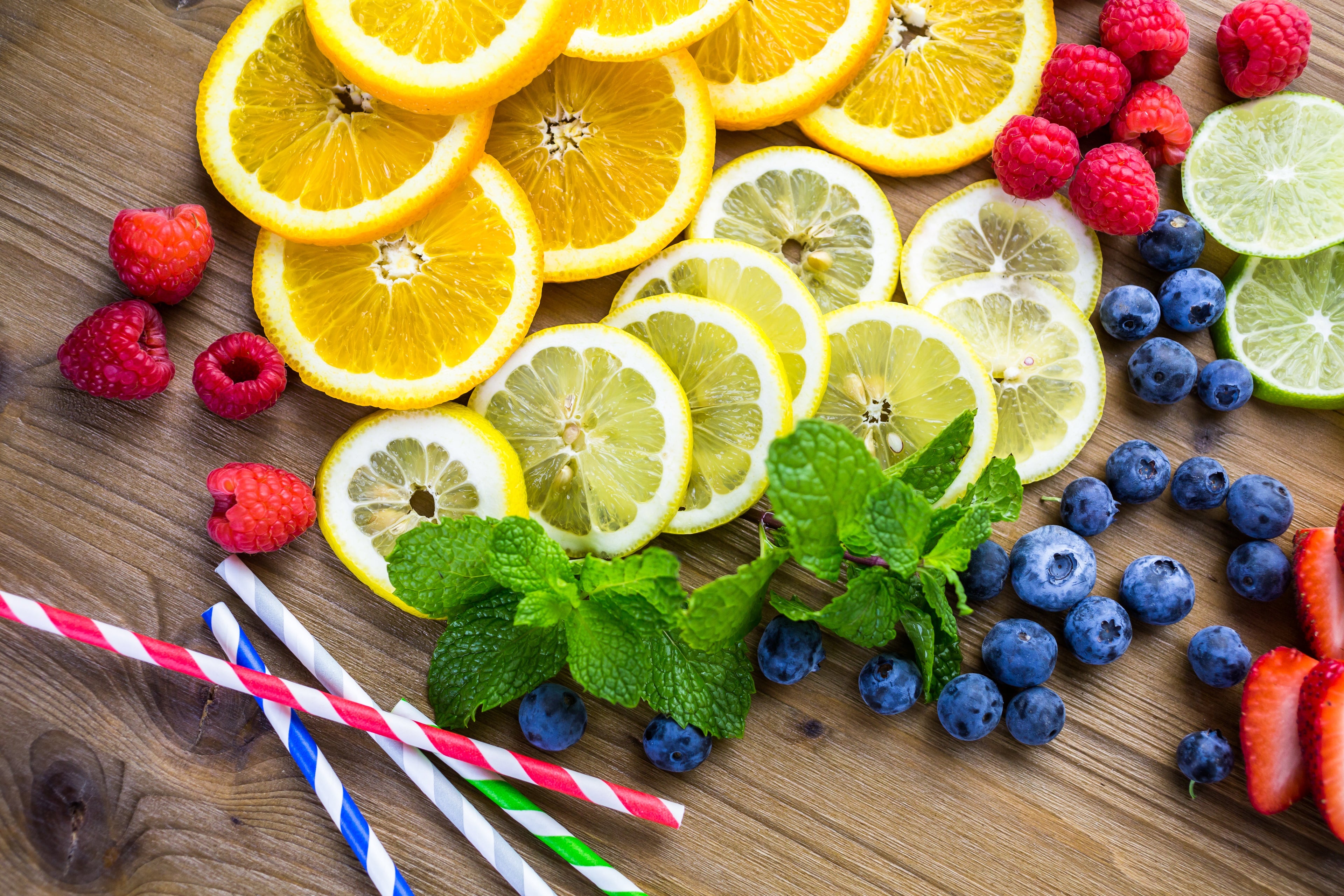 Wooden table with sliced lemons, oranges, blueberries, raspberries, strawberries, mint leaves and striped paper straws.