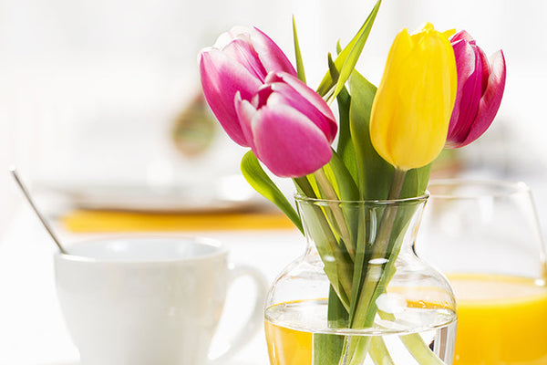 Bunch of tulips in a vase on a table with orange juice and a cup of coffee with a spoon in it.