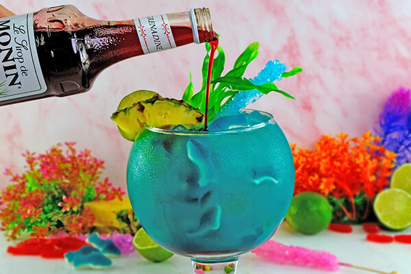 A large fishbowl glass filled with a bright blue cocktail, garnished with gummy sharks, pineapple wedge, lime, and green fronds. A bottle of Monin grenadine is being slowly poured into the drink to add a "blood in the water" effect"