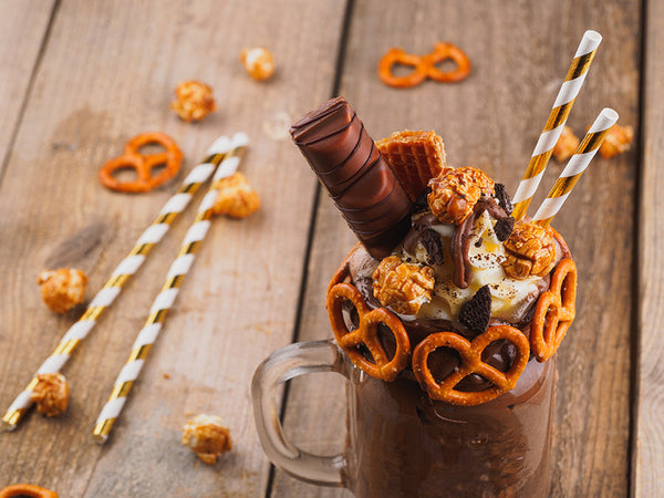 Chocolate milkshake garnished with a pretzeled rim, topped with whipped cream, caramel corn, cookie bits, cookies, and two gold striped straws.