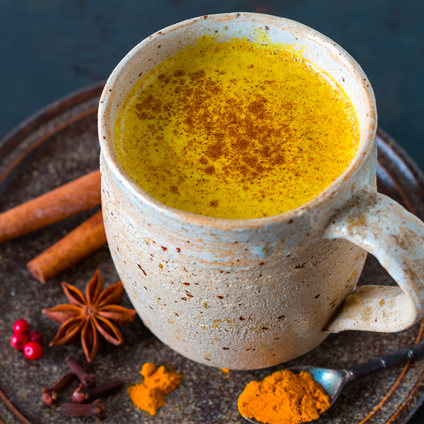 Turmeric drink image in a ceramic mug on a plate with cinnamon sticks and a spoonful of cinnamon.